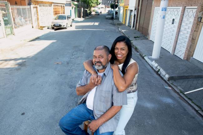 Imagem mostra pai e filha abraçados, com a filha por trás do pai. Eles seguram as mãos e estão no meio de uma rua residencial
