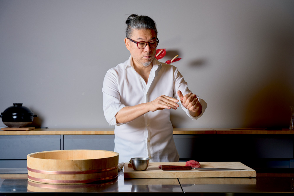 Sushiman de camisa social preparando niguiri em bancada de madeira.