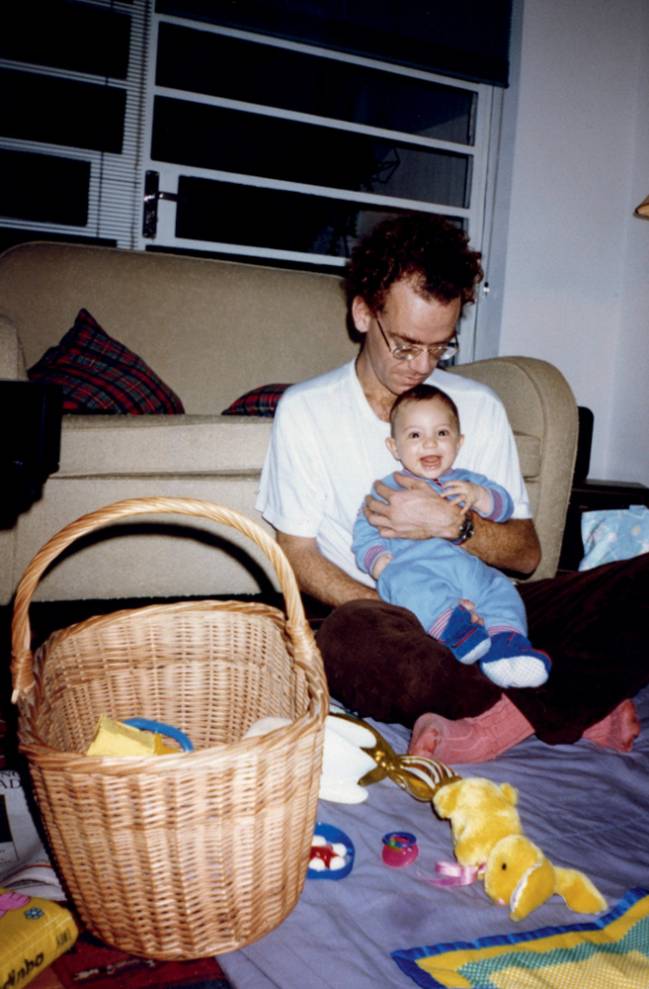 Foto antiga mostra Nando sentado em um tapete com o filho Sebastião, ainda bebê, no colo. Eles estão em uma sala