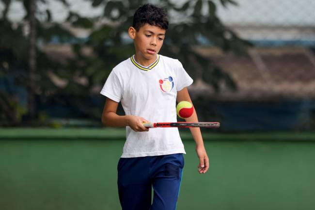 Imagem mostra menino com raquete de tênis na mão, batendo bola de tênis verticalmente em uma quadra