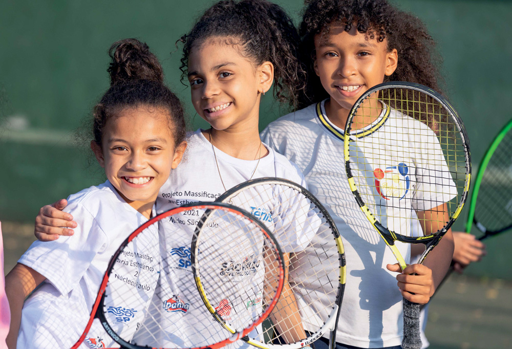 Imagem mostra três crianças sorridente mostrando raquetes de tênis