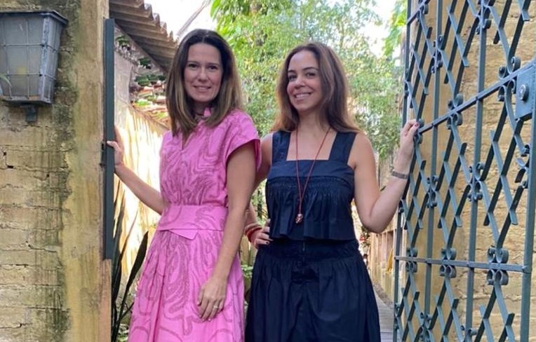 Duas mulheres posam de pé na entrada de uma casa. A da esquerda veste vestido rosa claro e a da direita um vestido com alças azul-escuro.