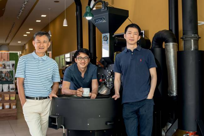 Imagem mostra pai e dois filhos em cafeteria, apoiados em equipamento grande de café. Eles estão em uma cafeteria, posicionados lado a lado e sorriem para a foto