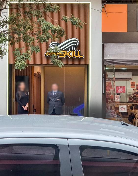 Imagem mostra fachada de loja durante o dia. Duas pessoas estão próximas da porta do local, uma mulher e um homem trajados com roupas sociais. Em cima, lê-se no letreiro: Arena Skill