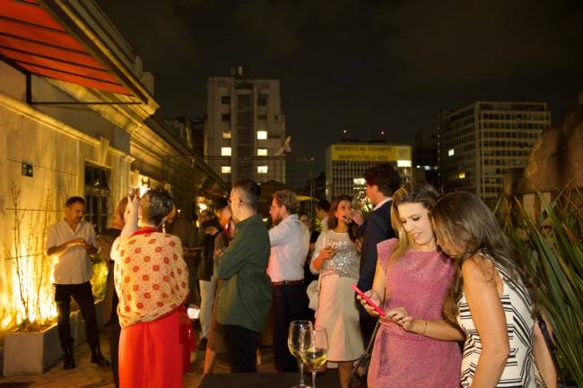 Imagem mostra várias pessoas conversando em terraço, durante a noite