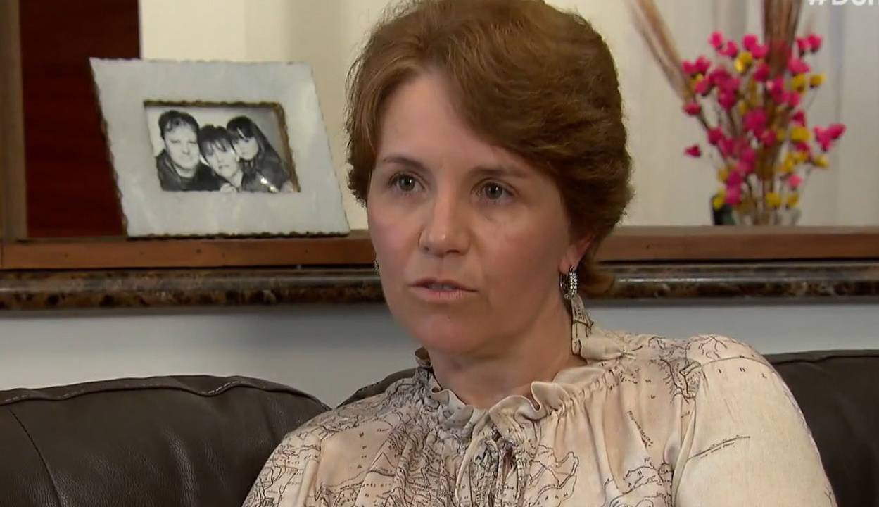 uma mulher branca de cabelos ruivos e curtos sentada em um sofá. atrás dela, um porta-retrato com uma foto da família.