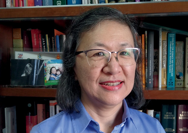 Mulher asiática com cerca de 60 anos sorri para câmera. Ela veste camisa social azul, óculos de grau. Está em frente a estante de livros.