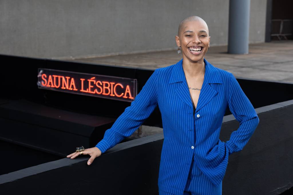 2 – Malu Avelar, Sauna Lésbica (Leo Martins) Mulher careca negra sorri em frente a letreiro que diz