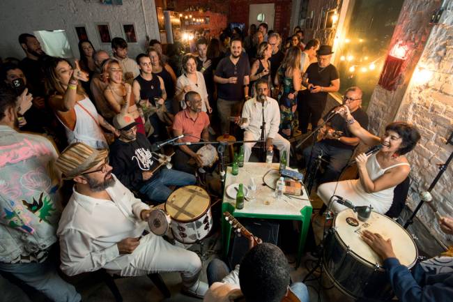 Imagem mostra roda de samba com homens tocando instrumentos e mulher cantando. Ambiente, fechado, está cheio de frequentadores de bar