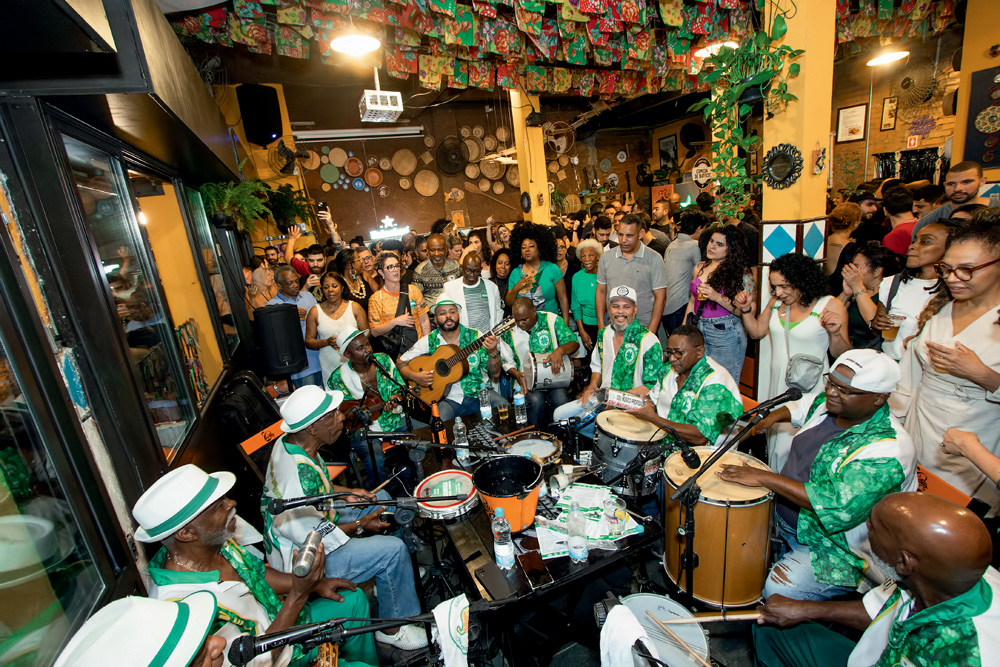Imagem mostra grande roda de samba com várias pessoas reunidas em um bar ao redor de grupo com homens, que estão sentados tocando diversos insturmentos