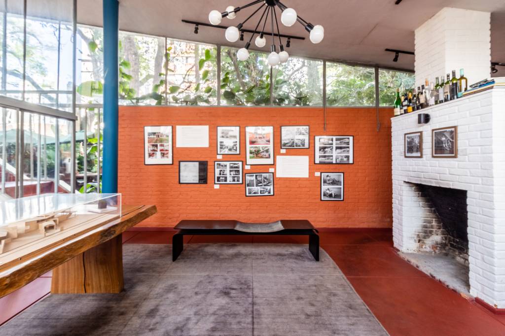 Parede laranja, teto alto, grande lustre, móveis de madeira, quadros e tapete cinza aparecem em destaque em sala de estar.