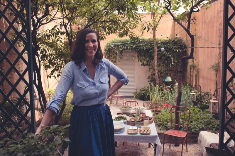 A imagem mostra Gabriela Barretto em um pequeno quintal com árvores e uma mesa montada ao fundo.