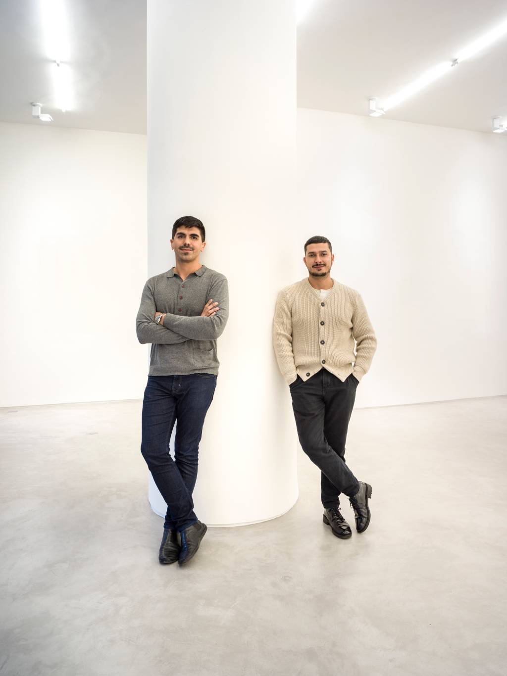 6 – Ian Duarte e Allann Seabra (Rômulo Fialdini-Divulgação) Dois homens brancos posam lado a lado apoiados em coluna branca. Ambos vestem calças pretas, o da esquerda com suéter cinza e o da direita cardigã claro.