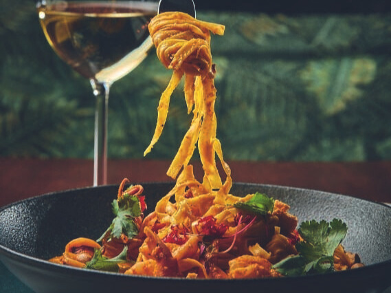 A imagem mostra uma mão segurando um garfo com macarrão enrolado nele. Embaixo um prato fundo com macarrão fettuccine e frutos do mar. Ao fundo, uma taça com vinho branco.