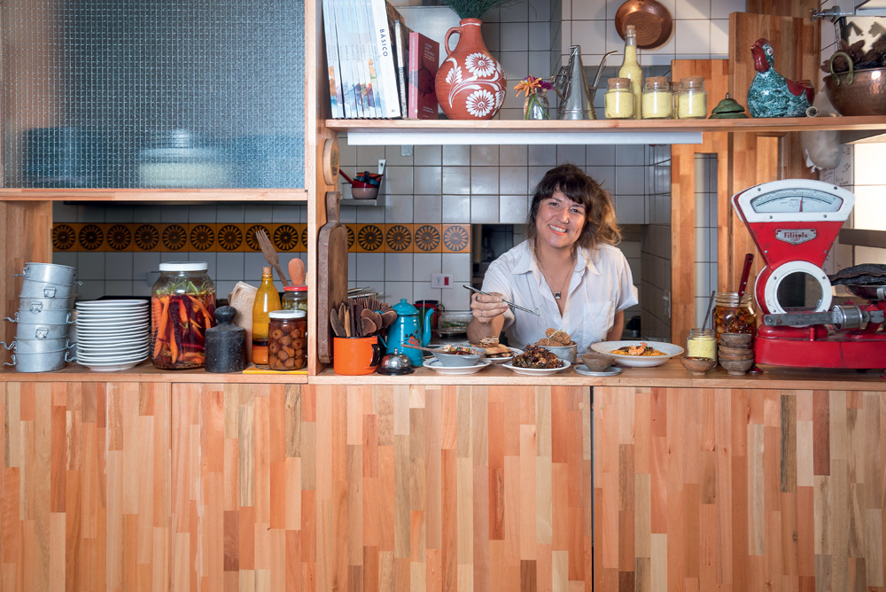 A imagem mostra uma mulher atrás de um balcão de madeira, ela sorri para a foto. No balcão a sua frente tem seis pratos com comidas. À esquerda no balcão tem uma pilha de pratos, panelas e potes de tempero.