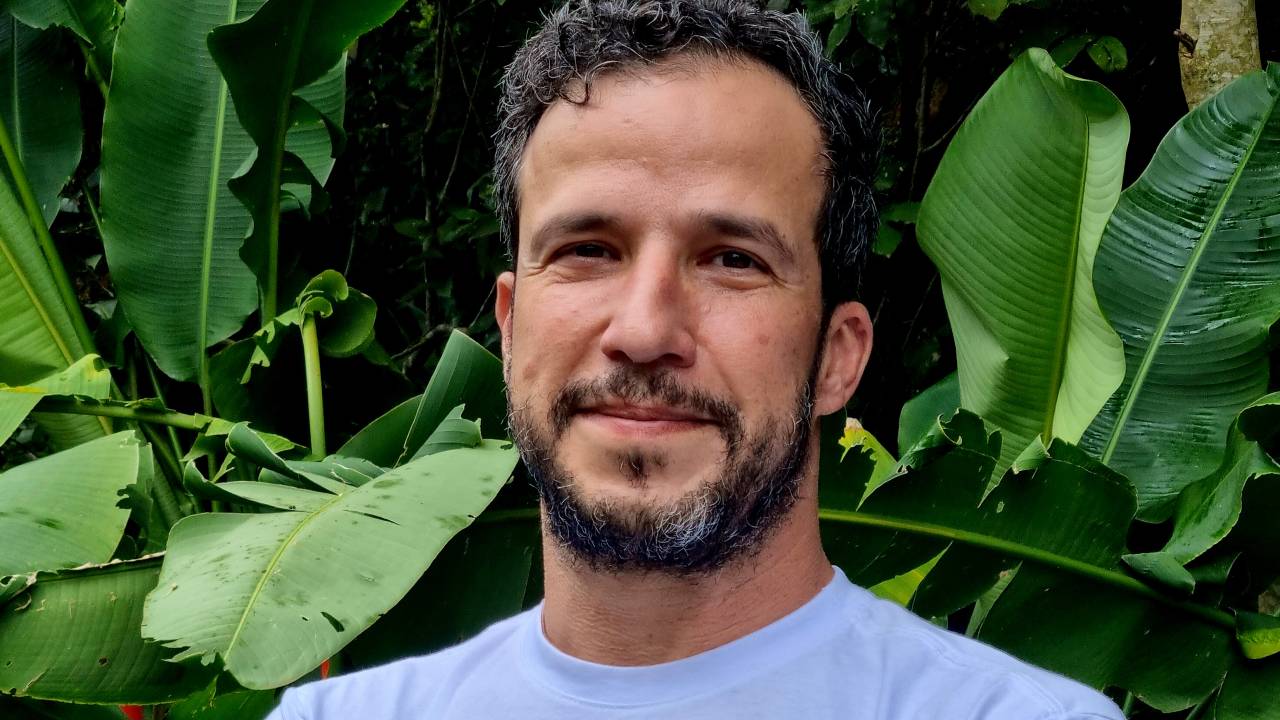 Homem branco de barba dá meio sorriso. Veste camiseta branca e posa em frente a área verde.