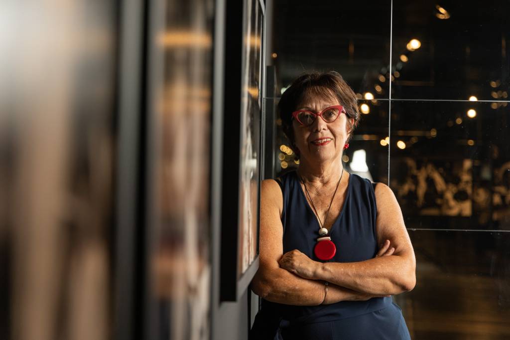 BIENAL2 Senhora de cabelos curtos posa encostada em parede com fotografias, veste colar vermelho e vestido azul-escuro. Usa óculos de grau.