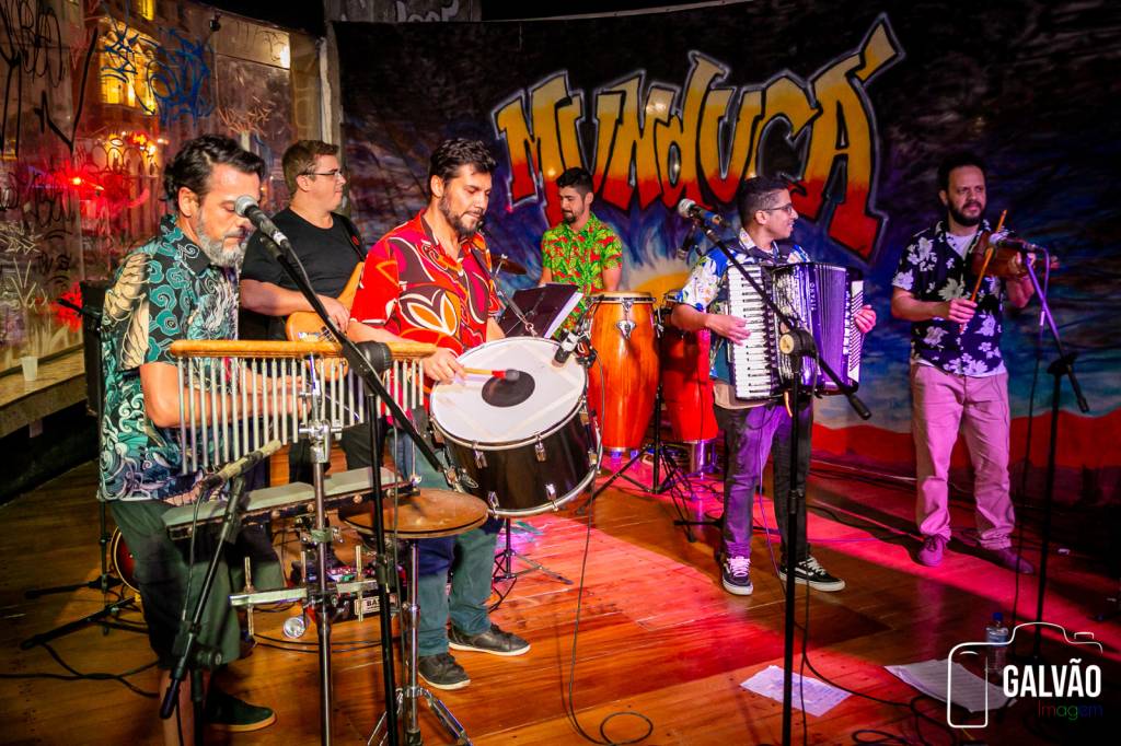 Foto iluminada em tom vermelho exibe integrantes de banda de forró manipulando seus instrumentos musicais.