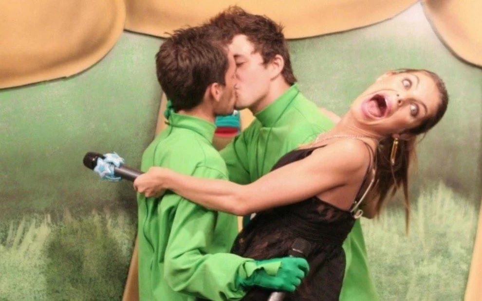 Imagem exibe dois homens de verde se beijando e apresentadora com cara de surpresa no meio.