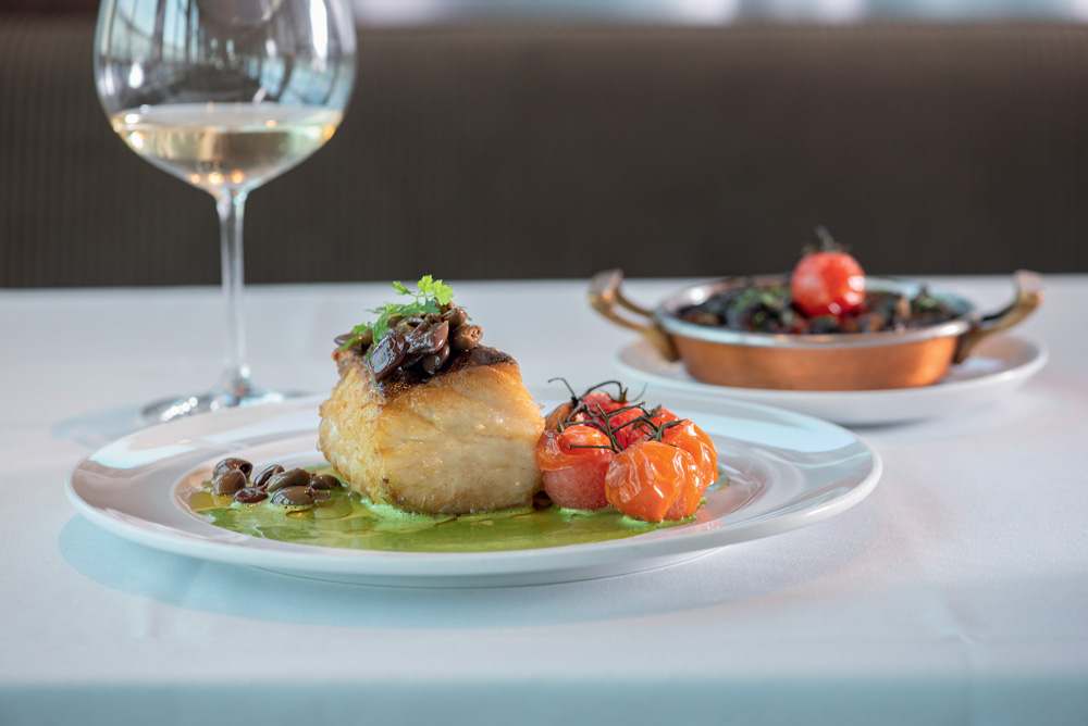 Prato branco com pedaço de bacalhau, legumes e molho de manjericão embaixo.