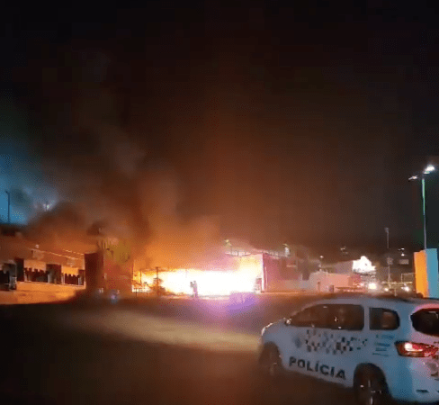 um estande pegando fogo ao fundo e uma viatura de polícia a frente