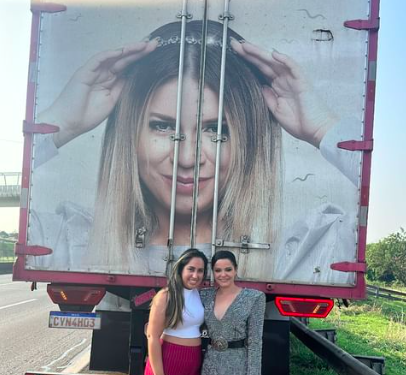 duas mulheres brancas abraçadas no acostamento de uma estrada e, ao fundo, a traseira de um caminhão com uma foto enorme da cantora marília mendonça