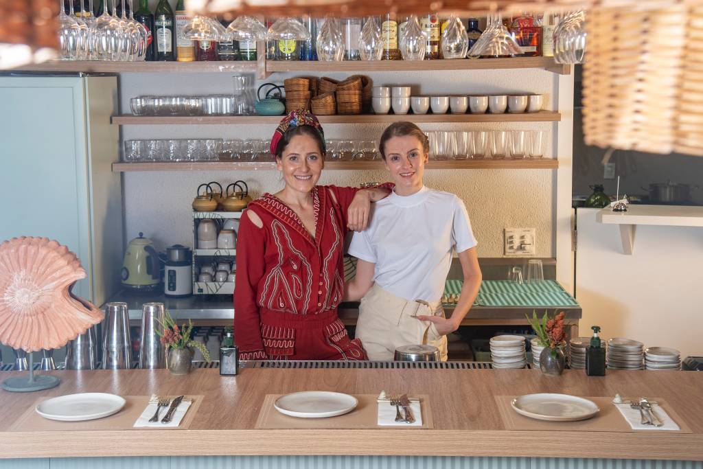Duas mulheres, uma de vermelho à esquerda e outra de branco e avental à direita atrás de balcão de madeira em restaurante