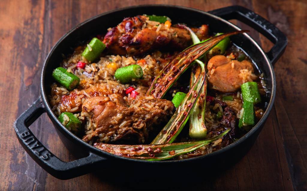 A imagem mostra uma panela preta em cima de uma mesa de madeira vista de cima. Dentro, uma mistura de arroz, com quiabos e pedaços de frango.