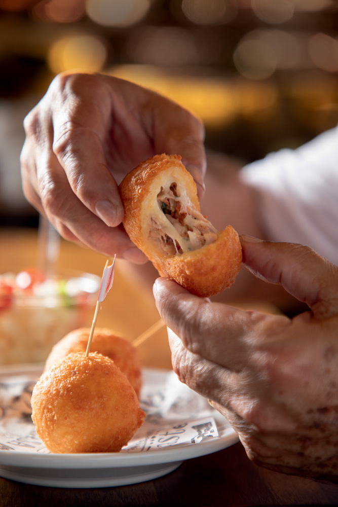 Duas mãos abrem um bolinho empanado, o recheio de queijo aparece.