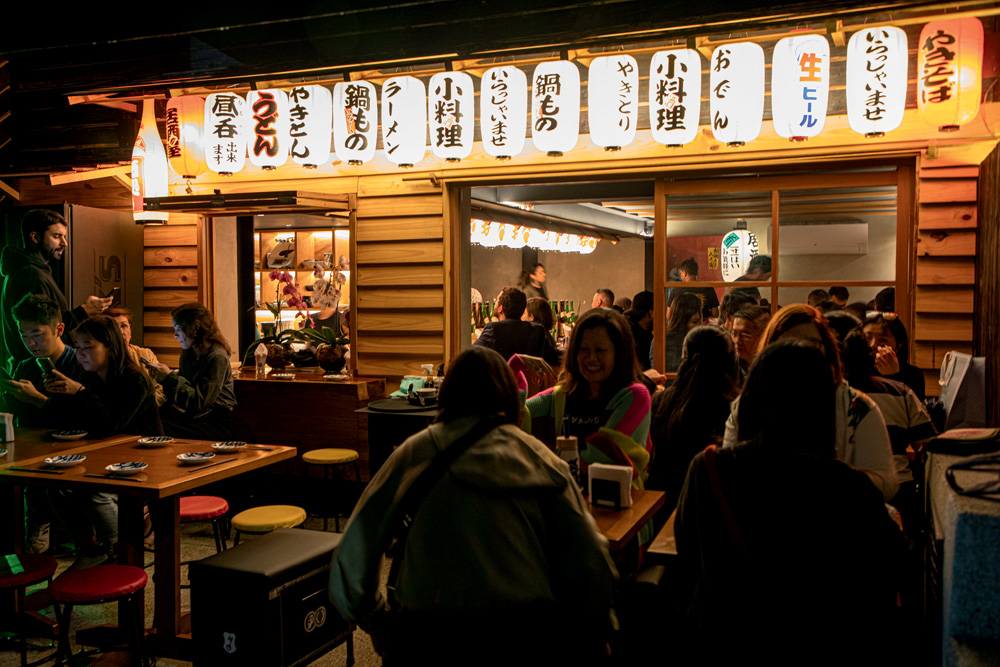 Fachada do restaurante com parede de tacos de madeira, em cima da porta várias lanternas brancas com ideogramas em japonês e coreano. Na frente da fachada, mesas de madeira e banquinhos coloridos com pessoas sentadas.