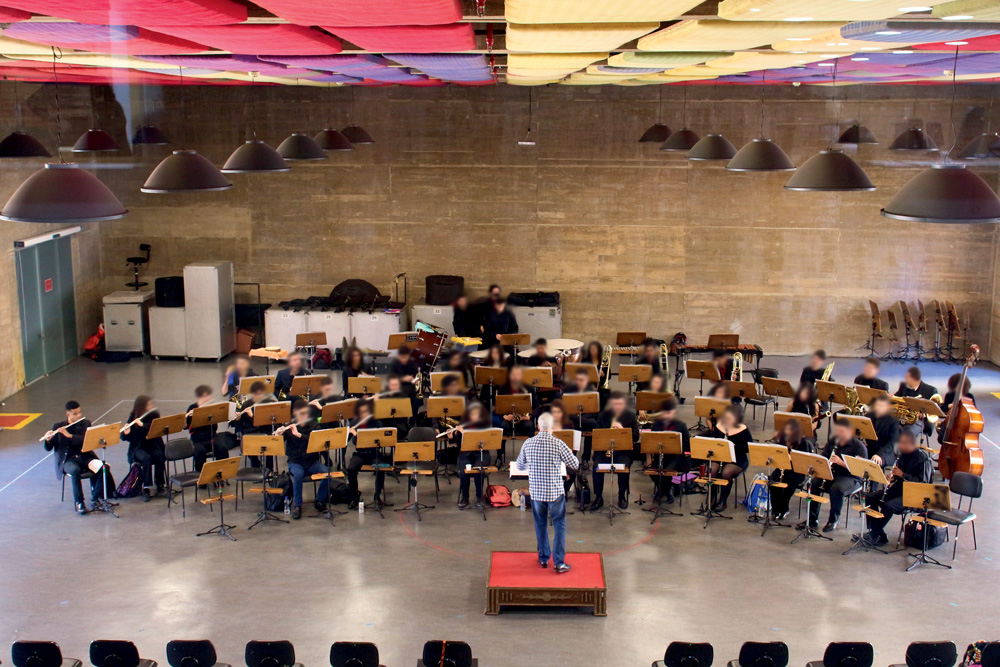 Imagem mostra orquestra ensaiando, com dezenas de jovens tocando instrumentos clássicos, com regente à frente do conjunto, em ambiente fechado e bem iluminado