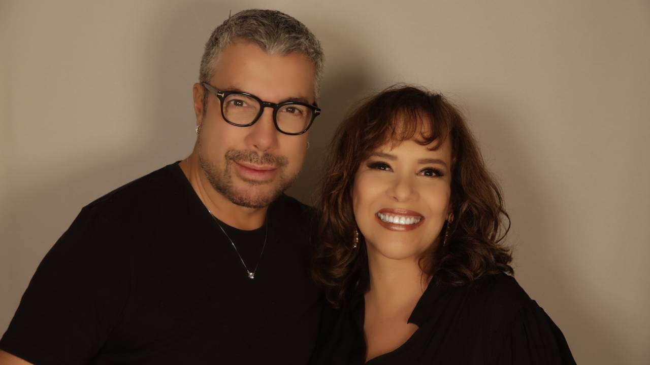 Homem grisalho de óculos de grau e mulher de franja e cabelo castanho sorriem e encaram a câmera, ambos vestindo roupas pretas.