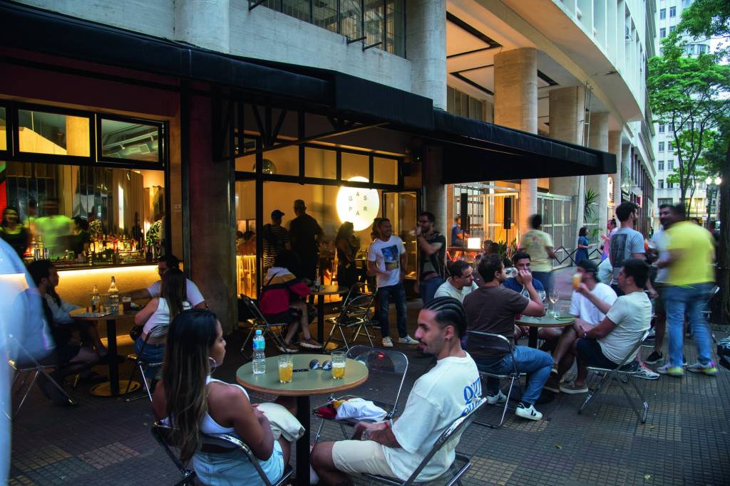 A fachada do bar, com mesas do lado de fora e pessoas sentadas, bebendo e conversando.