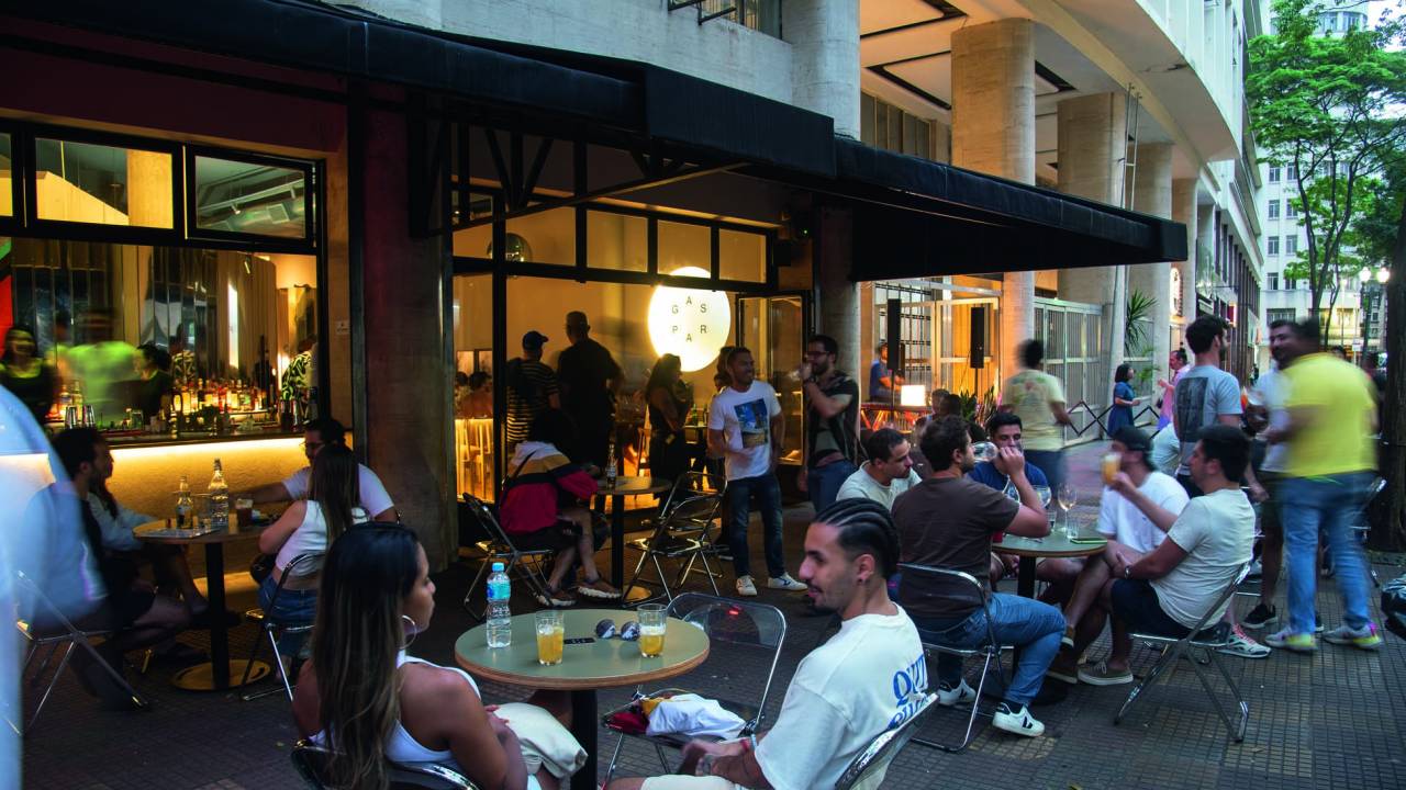 A fachada do bar, com mesas do lado de fora e pessoas sentadas, bebendo e conversando.