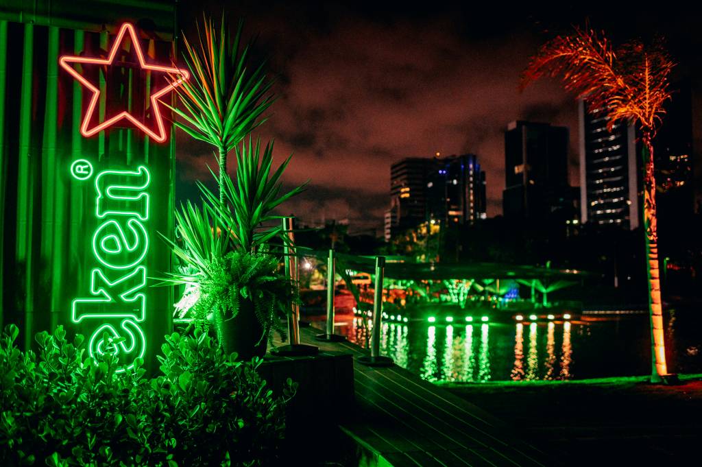 Imagem destaca bar em luz verde no meio de rio e prédios iluminados à noite de fundo.