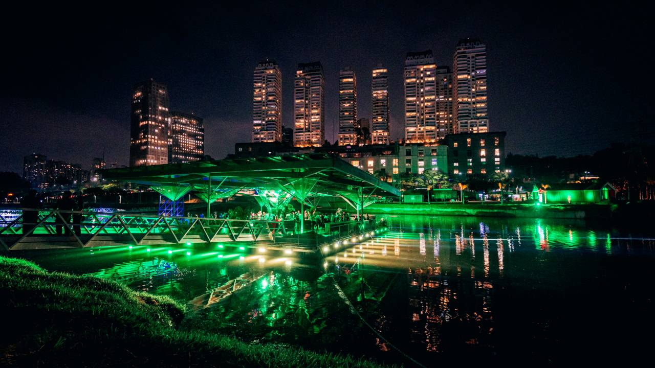 Imagem destaca bar em luz verde no meio de rio e prédios iluminados à noite de fundo.