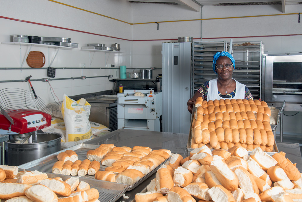 Mulher segurando uma bandeja lotada de pães sorri para a foto dentro de uma cozinha.