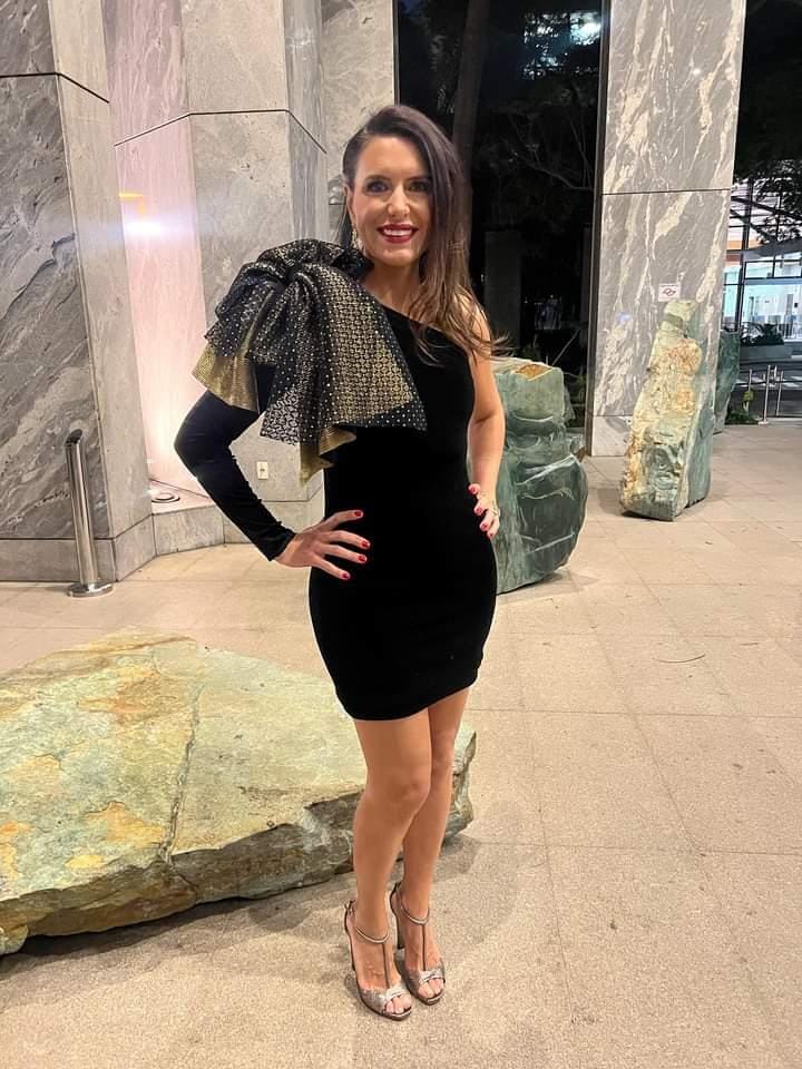 Juliana Meyer (Arquivo Pessoal) Mulher branca de cabelos castanhos lisos e longos posa em grande sala com vestido de festa preto, com bolinhas em detalhe em um dos ombros.