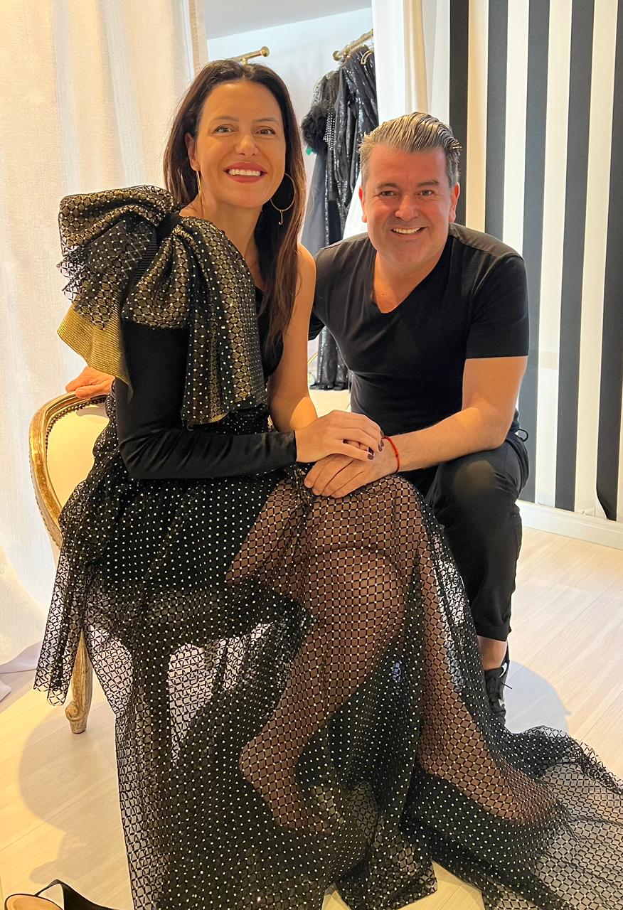 Juliana Meyer e Geraldo Couto (Arquivo Pessoal) (1) Mulher sentada em cadeira dourada sorri ao lado de homem grisalho de joelhos. Ela veste vestido preto com saia transparente e detalhes em um dos ombros. Ele veste camiseta e calça pretas.