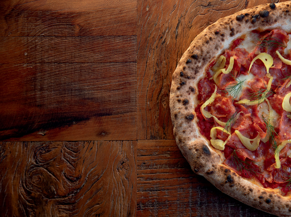 Imagem mostra metade de uma pizza com molho de tomate e pedaços de linguiça sobre um fundo de madeira.
