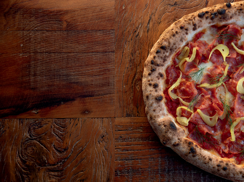 COMER & BEBER 2023 – La Crosta Forneria Imagem mostra metade de uma pizza com molho de tomate e pedaços de linguiça sobre um fundo de madeira.
