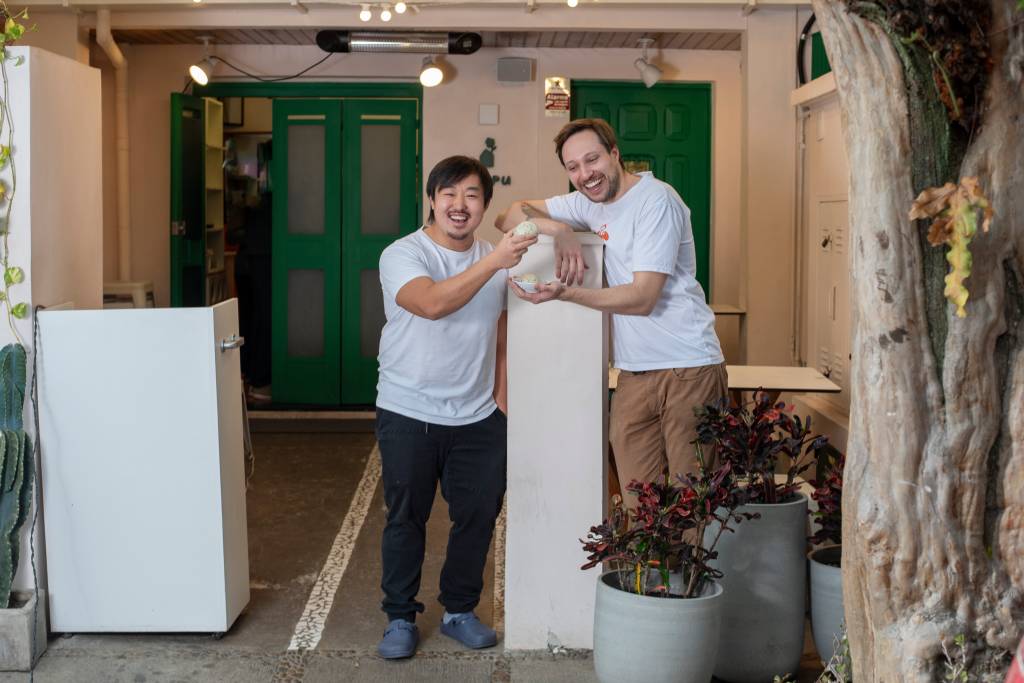 Dois homens na fachada de restaurante. À esquerda, homem asiático. À direita, homem branco.