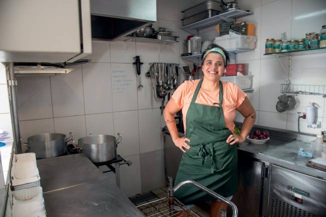 Mulher com avental verde com as mãos na cintura sorri para a foto. Ela está dentro de uma cozinha.