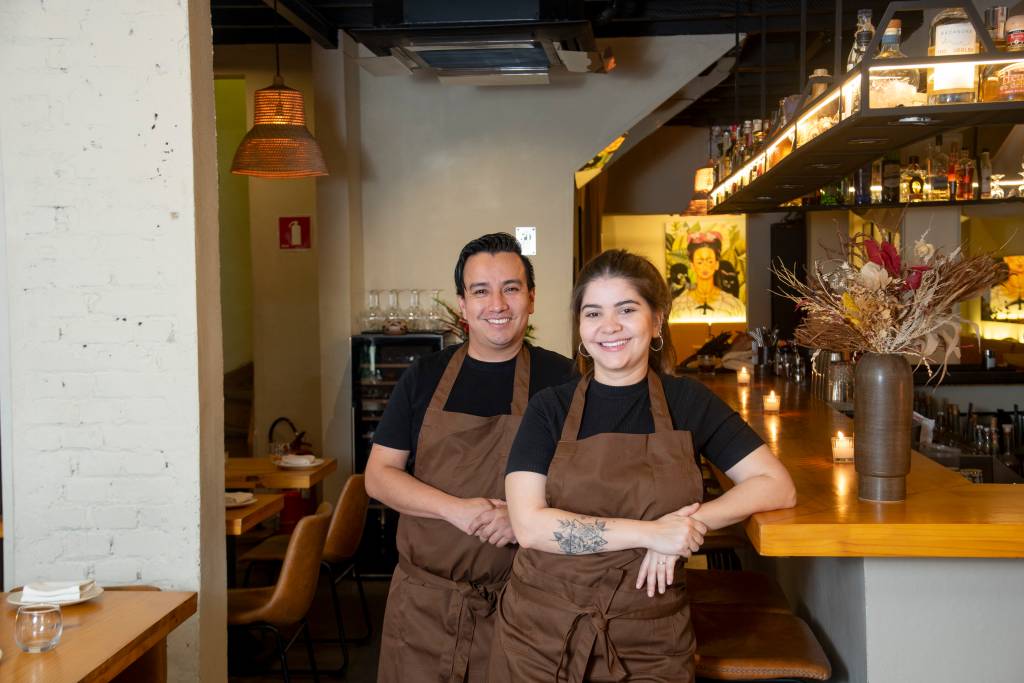 Homem, à esquerda, e mulher, à direita, de camiseta preta e avental marrom em salão de restaurante.