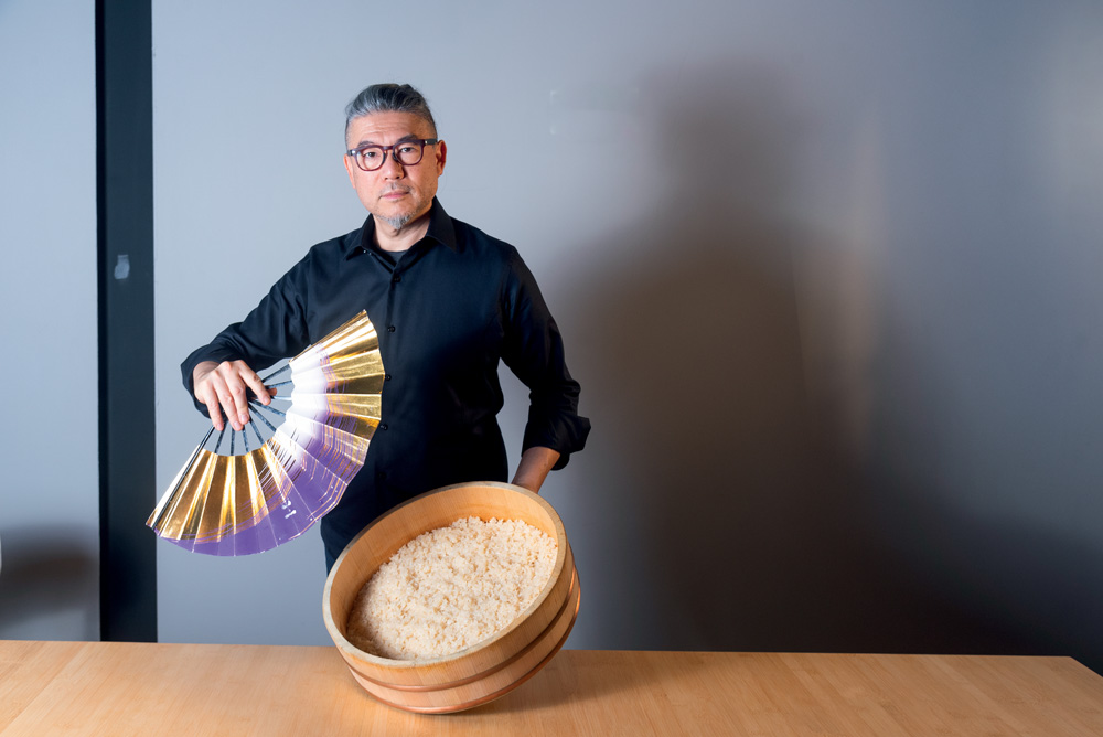 Um homem segura um leque em uma mão e uma bacia de arroz em outra.