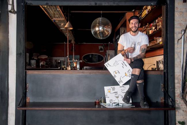 Homem de camiseta branca e boné preto sentado na bancada de um bar, ele sorri para a foto. Em uma mão segura um copo e na outra uma folha de papel com desenhos de drinques. Ao fundo é possível ver as prateleiras do bar com garrafas de bebida.