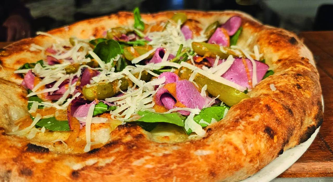 A imagem mostra uma pizza vista de cima, com as bordas bem grossas e fatias de pastrami, rúcula e queijo.