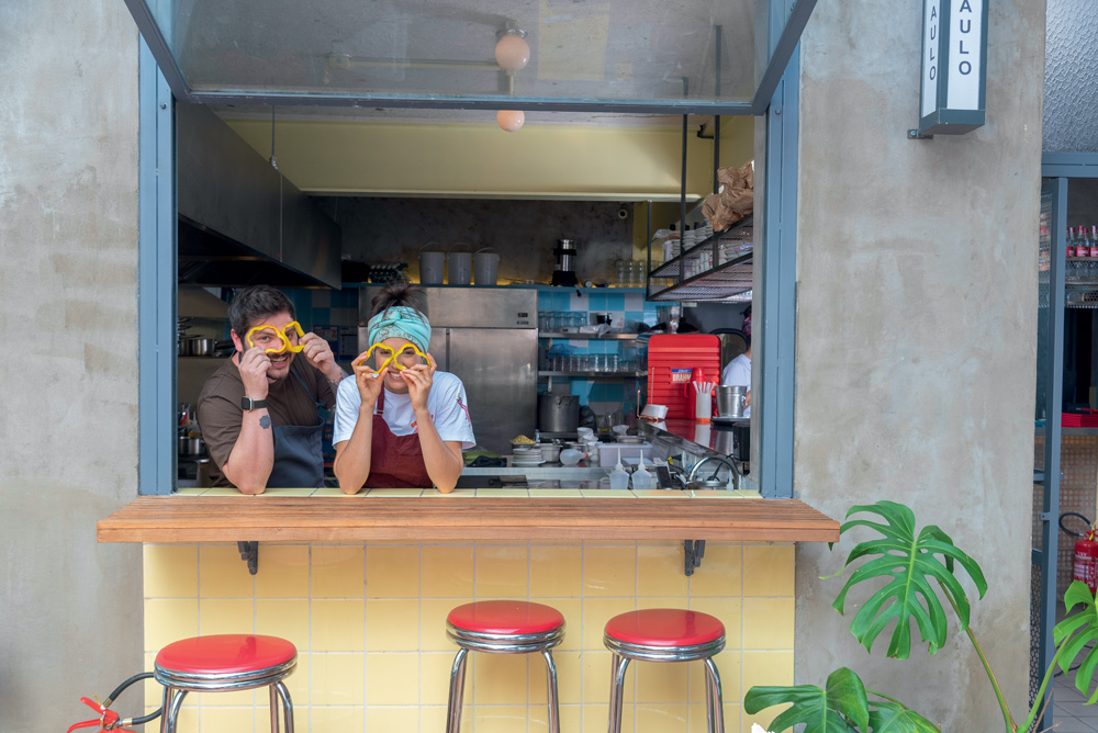 Júlia e Gabriel atrás do balcão de azulejos amarelos do restaurante, seguram duas rodelas amarelas cada um sobre seus olhos.