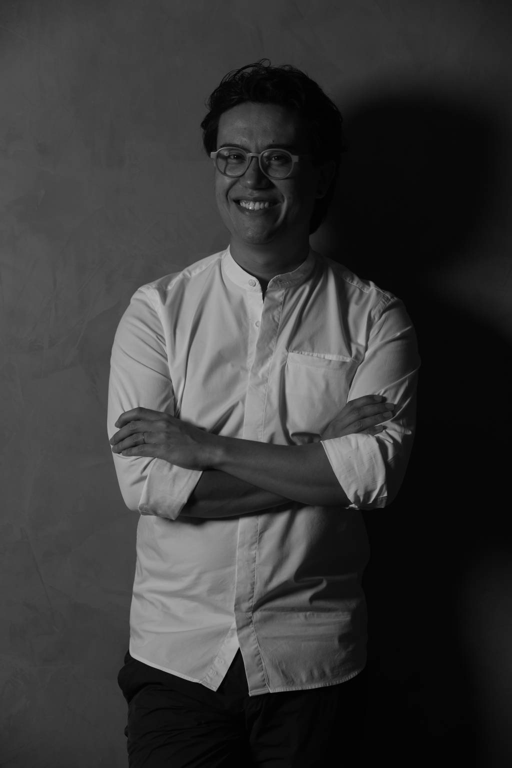 Foto em preto e branco de homem com traços levemente asiáticos, com camiseta longa branca, de braços cruzados.