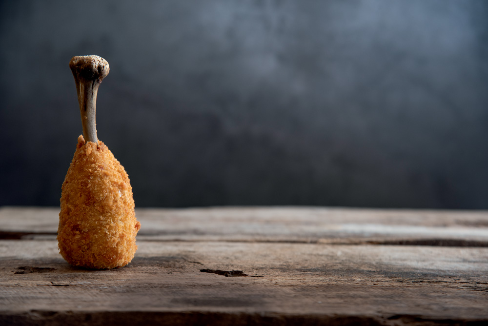 Uma coxinha com um osso em cima sobre uma mesa de madeira. O fundo é uma parede preta.